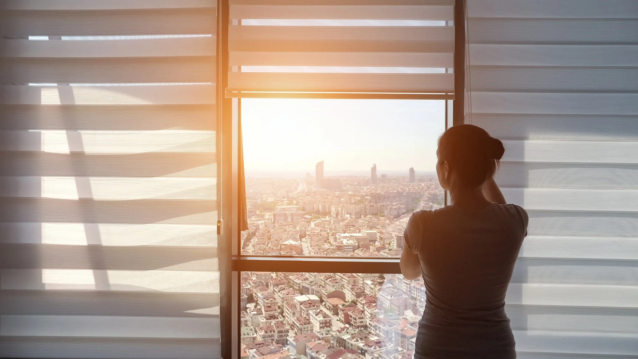 Person standing beside a large window with sunlight and a city view